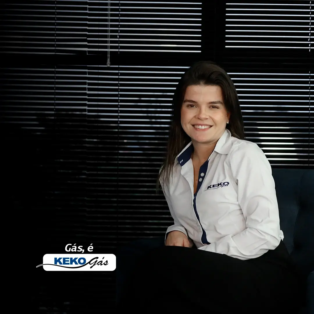 Imagem de uma mulher jovem, caucasiana, vestindo uma camisa social branca com a logomarca da keko gás, sentada em uma poltrona em frente a uma janela de escritório